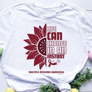May include: White t-shirt with a maroon sunflower design and the text "Life Can Change In An Instant" with a maroon ribbon below. The text "Multiple Myeloma Awareness" is printed below the ribbon.