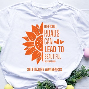 Pode incluir: Camiseta branca com um gráfico de girassol laranja e o texto "Difficult Roads Can Lead To Beautiful Destinations" e "Self Injury Awareness".