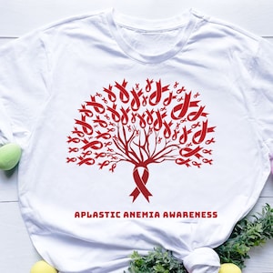 Può includere: Maglietta bianca con un design a forma di albero di nastro rosso e il testo "Aplastic Anemia Awareness".