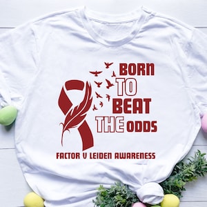 May include: White t-shirt with a maroon ribbon and feather design, accompanied by the text "BORN TO BEAT THE ODDS" and "FACTOR V LEIDEN AWARENESS". The design includes flying birds, creating a symbolic and motivational message.