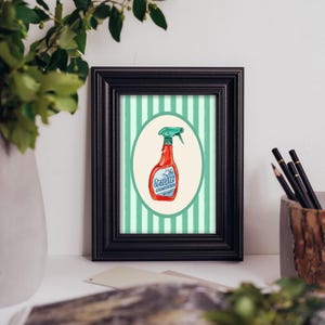 May include: A framed print featuring a red spray bottle with a green nozzle centered within an oval frame. The background consists of vertical light green and white stripes. The bottle label displays the text Opalite in blue lettering.