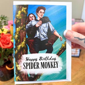 Op de afbeelding: Witte wenskaart met een kleurrijke illustratie van twee mensen op een boomtak. De kaart heeft de tekst "Happy Birthday SPIDER MONKEY" in zwart en wit. De achtergrond is een blauwe lucht.