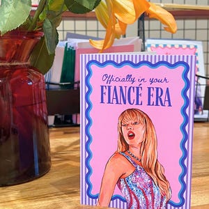 May include: A greeting card with the text "Officially in your FIANCÉ ERA" and an illustration of a woman in a colorful dress. The card has a pink background with a blue and white striped border. A red vase with flowers is in the background.