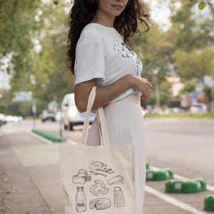 Op de afbeelding: Een cr&egrave;mekleurige canvas draagtas met een zwarte lijntekening van diverse zuivelproducten, waaronder een melkfles met het woord Milk erop, een kaasrasp en verschillende soorten kaas in de vorm van stukken en hele kazen.