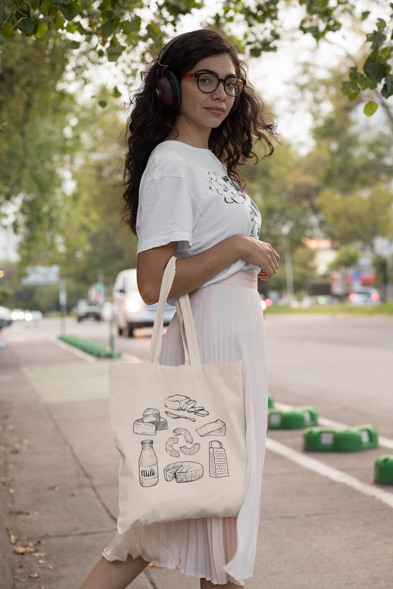 Op de afbeelding: Een cr&egrave;mekleurige canvas draagtas met een zwarte lijntekening van diverse zuivelproducten, waaronder een melkfles met het woord Milk erop, een kaasrasp en verschillende soorten kaas in de vorm van stukken en hele kazen.