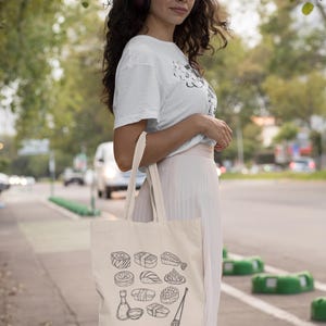 A person wearing headphones, glasses, a white t-shirt, and a light skirt, stands outdoors holding a cream canvas tote bag with a black line art sushi graphic.