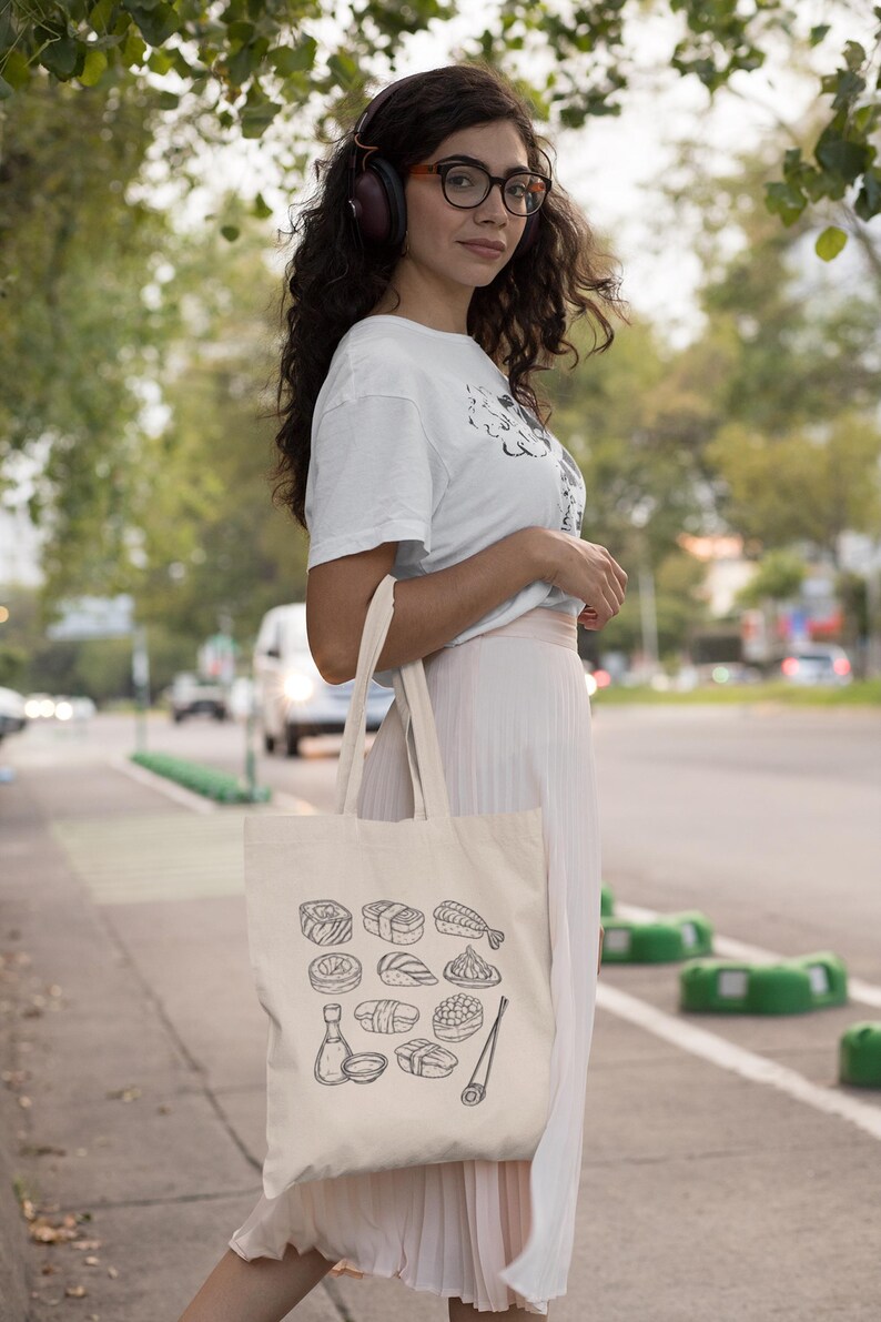 A person wearing headphones, glasses, a white t-shirt, and a light skirt, stands outdoors holding a cream canvas tote bag with a black line art sushi graphic.