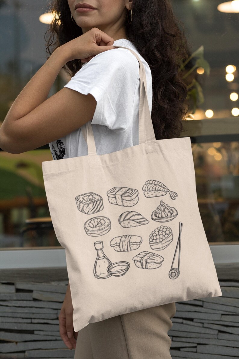 A person with long dark hair, wearing a white t-shirt, holds a cream canvas tote bag with a black line art sushi graphic over their shoulder.