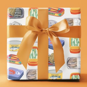 Puede incluir: Una caja de regalo blanca con una cinta dorada. La caja está envuelta en papel con un patrón de latas de sardinas y caballa coloridas. El texto en las latas dice "Sardines" y "Caballa".