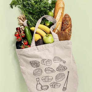 A cream canvas tote bag with a black line art sushi graphic is filled with groceries including greens, bread, and various fruits, against a light green background.