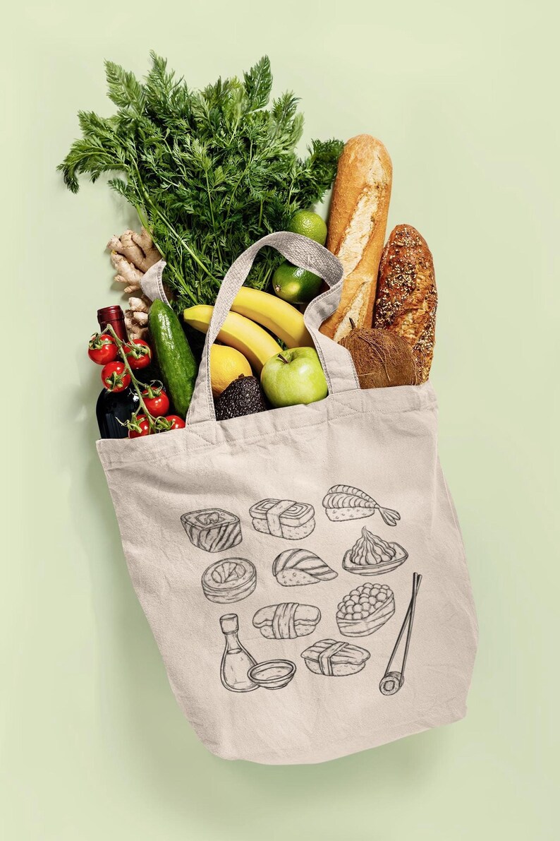 A cream canvas tote bag with a black line art sushi graphic is filled with groceries including greens, bread, and various fruits, against a light green background.