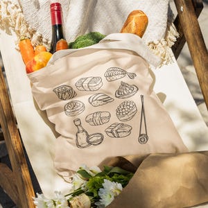 A cream canvas tote bag with a black line art sushi graphic sits in a wooden chair, filled with groceries including a bottle of wine, a baguette, and vegetables.