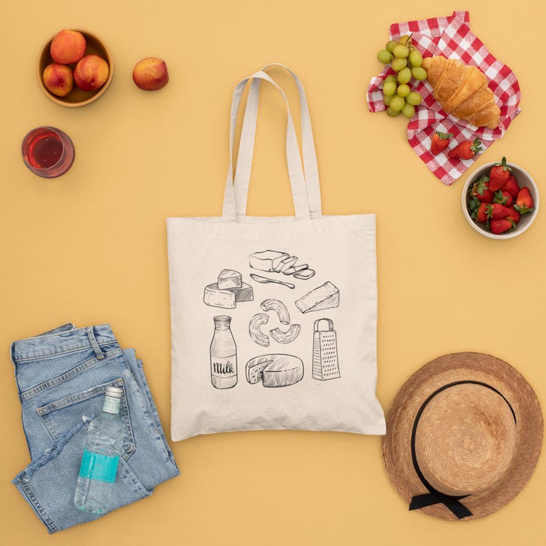 Op de afbeelding: Een beige canvas draagtas met een zwarte lijntekening van diverse zuivelproducten, waaronder een melkfles, een kaasrasp en verschillende soorten kaas. De tas is omringd door perziken, een croissant, druiven, aardbeien, een spijkerbroek en een hoed.