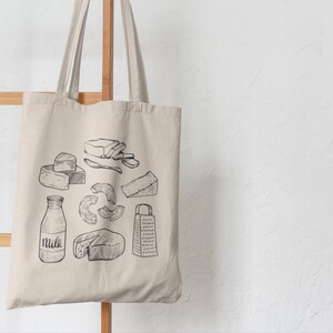 Op de afbeelding: Een beige canvas draagtas met een zwarte lijntekening van diverse zuivelproducten, waaronder een melkfles met het woord Milk erop, een kaasrasp en verschillende soorten kaasstukken die op de voorzijde van de tas zijn afgebeeld.