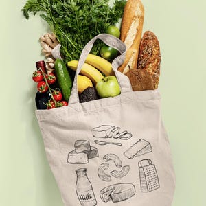 Op de afbeelding: Een beige canvas tas met zwarte lijntekeningen van zuivelproducten, een rasp en een fles met het label milk. De tas zit vol met verse producten, waaronder wortelen, bananen, een groene appel, een komkommer, gember en twee broden.