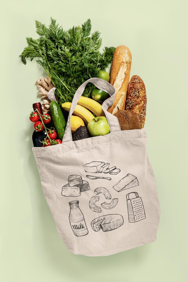 Op de afbeelding: Een beige canvas tas met zwarte lijntekeningen van zuivelproducten, een rasp en een fles met het label milk. De tas zit vol met verse producten, waaronder wortelen, bananen, een groene appel, een komkommer, gember en twee broden.