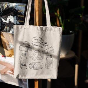 Op de afbeelding: Een cr&egrave;mekleurige canvas draagtas met een zwarte lijntekening van diverse zuivelproducten, waaronder een melkfles met het opschrift Milk, een kaasrasp en verschillende soorten kaasstukken die in een cirkelvormig patroon zijn gerangschikt.