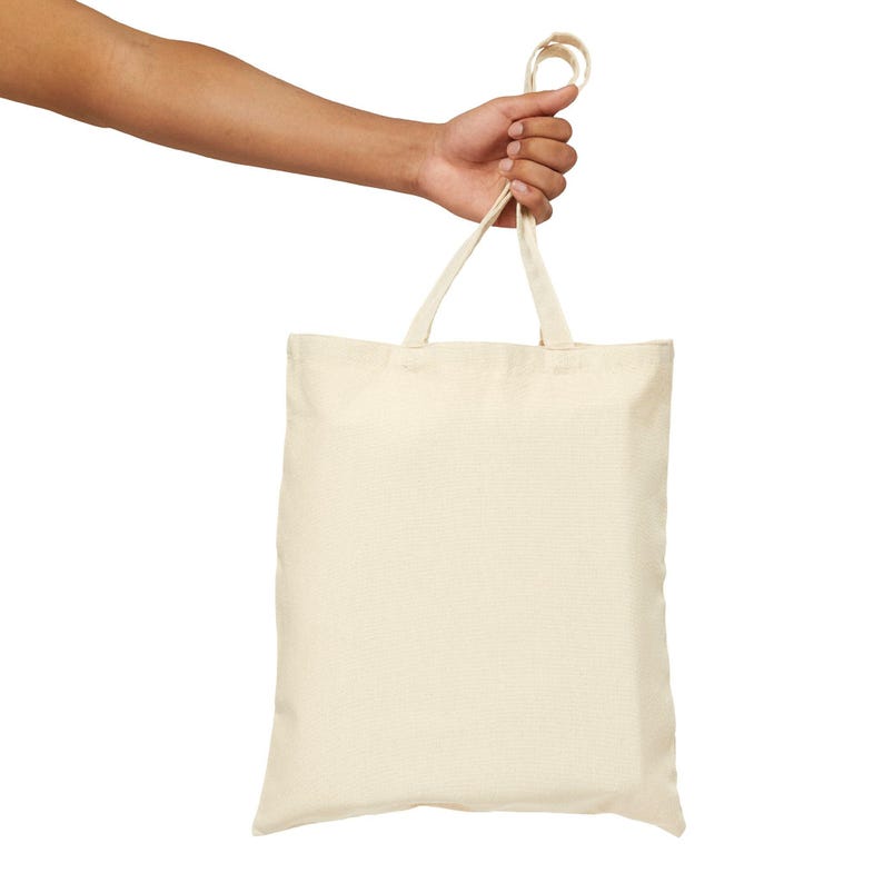 A person's arm and hand hold a blank, cream-colored canvas tote bag by its handles against a white background.