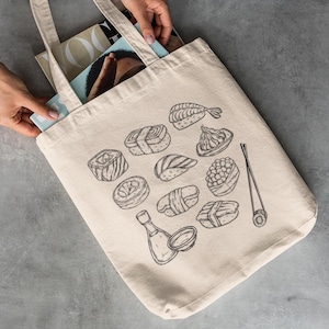 A pair of hands places books into a cream canvas tote bag with a black line art sushi graphic on a gray concrete surface.
