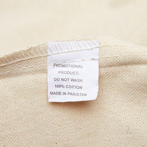 A close-up of a white fabric tag sewn into a cream-colored canvas bag, reading "PROMOTIONAL PRODUCT- DO NOT WASH 100% COTTON MADE IN PAKISTAN."