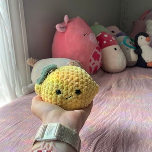 May include: A yellow crocheted lemon plush toy with a green stem and a smiling face. The toy is being held in a hand with several bracelets on the wrist.