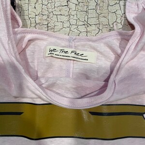 May include: A pink sleeveless shirt with a gold and black graphic print. The shirt has a tag that reads "We The Free" and "Large" with the text "Made in Indonesia / Fabrique en Indonesie".
