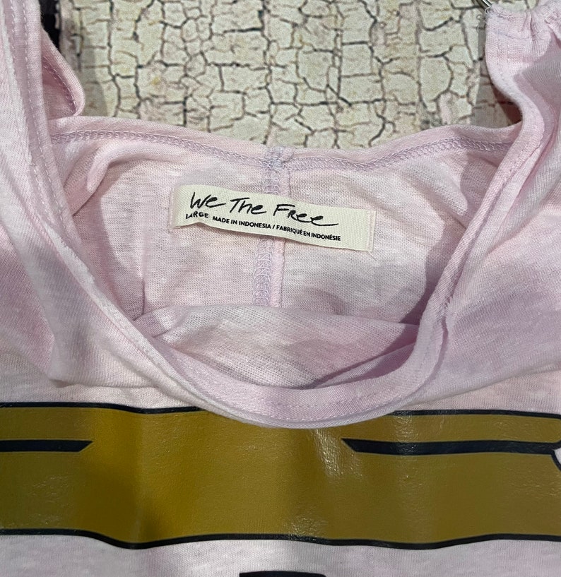 May include: A pink sleeveless shirt with a gold and black graphic print. The shirt has a tag that reads "We The Free" and "Large" with the text "Made in Indonesia / Fabrique en Indonesie".