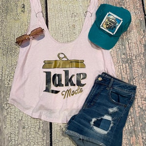 May include: Pink tank top with a gold can graphic and the text "Lake Mode". A teal baseball cap with the text "Tanned and Tipsy" is on the right side of the image. A pair of brown sunglasses and blue denim shorts are also included.