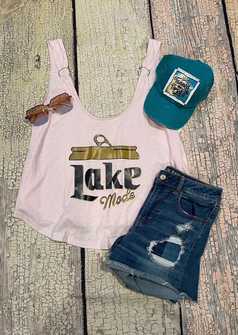 May include: Pink tank top with a gold can graphic and the text "Lake Mode". A teal baseball cap with the text "Tanned and Tipsy" is on the right side of the image. A pair of brown sunglasses and blue denim shorts are also included.