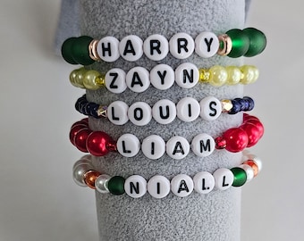 Könnte beinhalten: Fünf Perlenarmbänder mit den Namen "Harry", "Zayn", "Louis", "Liam" und "Niall", die in weißen Buchstaben ausgeschrieben sind.