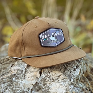 May include: A brown hat with a patch that reads "HAT DOWN Waterfowl" featuring a duck graphic. The hat has a curved brim, a green rope detail, and a white underside. The hat is resting on a rock.