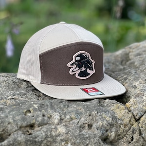Op de afbeelding: Een bruine en beige trucker-pet met een zwart-witte patch van een cartoon eend die een hoed draagt. De patch zegt "HOW".