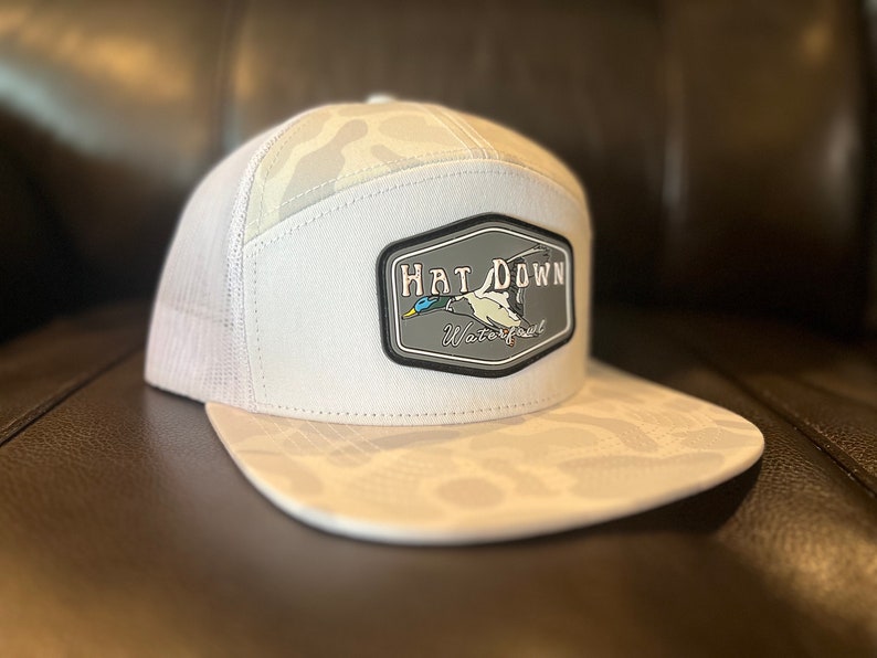 May include: White camouflage trucker hat with a black and grey patch that says "Hat Down Waterfowl" and features a flying duck.