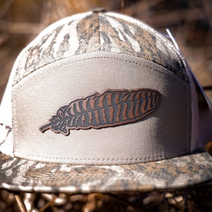 Puede incluir: Una gorra de b&eacute;isbol con estampado de camuflaje y un parche de cuero marr&oacute;n en forma de pluma en la parte delantera.