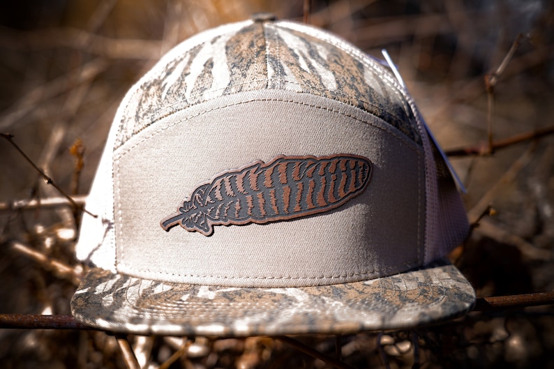 Puede incluir: Una gorra de b&eacute;isbol con estampado de camuflaje y un parche de cuero marr&oacute;n en forma de pluma en la parte delantera.