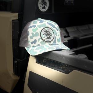 May include: A trucker hat with a camouflage pattern in shades of blue and white. The hat features a gray mesh back and a circular patch with the text "HAT DOWN WATERFOWL" and a duck graphic.