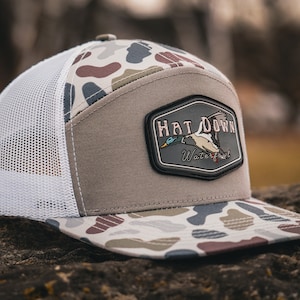 May include: A camouflage patterned trucker hat with a brown mesh back. The hat has a patch on the front that says "Hat Down Waterfowl" with a duck flying above the text.