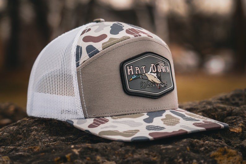 May include: A camouflage patterned trucker hat with a brown mesh back. The hat has a patch on the front that says "Hat Down Waterfowl" with a duck flying above the text.