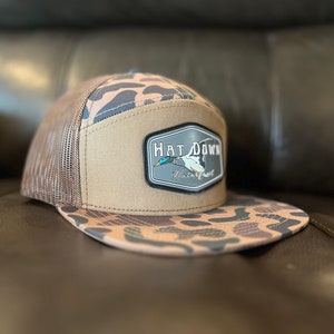 May include: A brown and tan camouflage trucker hat with a brown mesh back. The hat has a patch on the front that says "Hat Down Waterfowl".