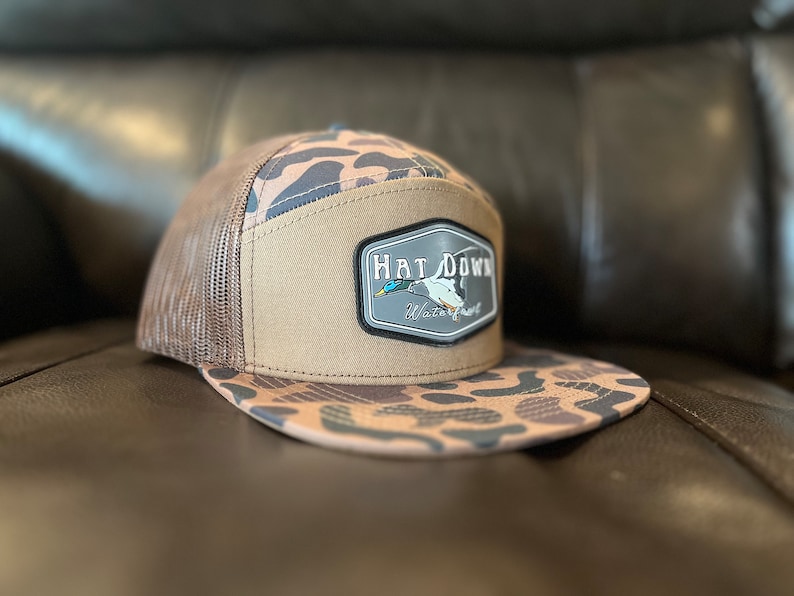 May include: A brown and tan camouflage trucker hat with a brown mesh back. The hat has a patch on the front that says "Hat Down Waterfowl".