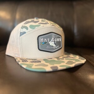 May include: A beige and green camouflage trucker hat with a black and white patch that says "Hat Down Waterfowl" and features a duck in flight.