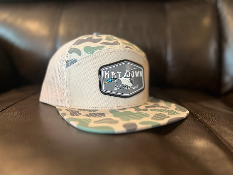 May include: A beige and green camouflage trucker hat with a black and white patch that says "Hat Down Waterfowl" and features a duck in flight.