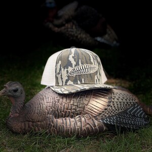 May include: A camouflage-patterned baseball cap with a white mesh back and a feather logo rests on a turkey decoy. The decoy is brown and tan, positioned on green grass. Another turkey is blurred in the background.