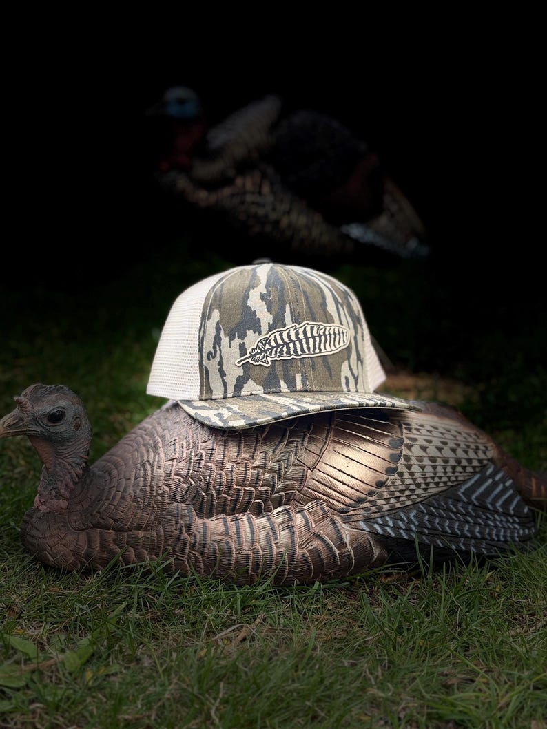 May include: A camouflage-patterned baseball cap with a white mesh back and a feather logo rests on a turkey decoy. The decoy is brown and tan, positioned on green grass. Another turkey is blurred in the background.