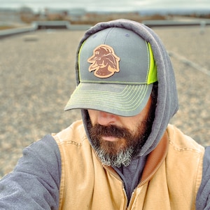 May include: A gray and neon green trucker hat with a brown leather patch featuring a duck design. The hat has a curved brim and a mesh back. The person in the image is wearing a gray hoodie and a tan vest.