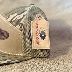 Puede incluir: Una gorra de camionero de camuflaje marr&oacute;n y verde con un parche de cuero marr&oacute;n y una etiqueta marr&oacute;n que dice "Mossy Oak" en letras blancas.