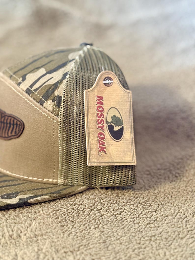 Puede incluir: Una gorra de camionero de camuflaje marr&oacute;n y verde con un parche de cuero marr&oacute;n y una etiqueta marr&oacute;n que dice "Mossy Oak" en letras blancas.