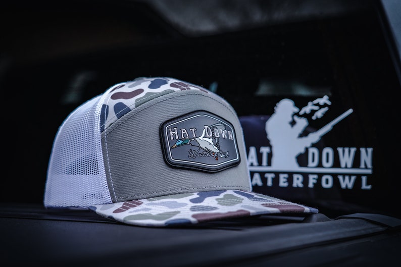 May include: A grey and white camouflage baseball cap with a black patch that says "Hat Down Waterfowl" and features a flying duck.