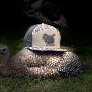 May include: A camouflage-patterned baseball cap with a mesh back and a tan front panel featuring a turkey silhouette. The hat rests on a turkey decoy in a grassy setting, with another turkey visible in the background.
