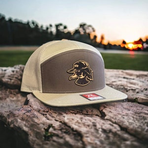 May include: A tan and brown trucker hat with a duck logo wearing a hat. The hat has a mesh back and a flat brim. The duck logo is brown and gold. The hat is sitting on a weathered log.